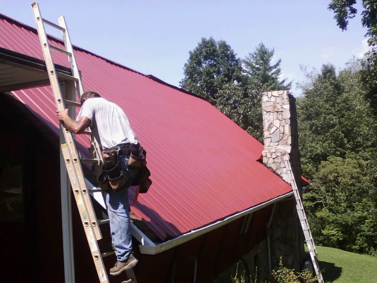 Roofer on a ladder beside a red metal roof and stone chimney — Soffit & Fascia Repair by Roofing Brothers in Luebbering